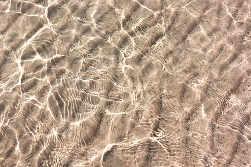 White sand pattern under clear transparent water with reflection of the sun in small waves from paradise beach