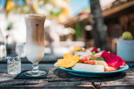 A glass of iced coffee and a fruit salad at a beach bar with a blurred beach background.