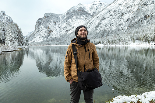 Young bearded man posing and looking away with Toblacher See (Dobbiaco Lake) on background in a snowy winter day. Reflections of mountains and trees on water; Dolomites, Italy