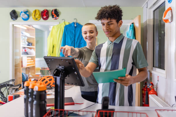 Teaching the Apprentice A front-view shot of a young apprentice salesman and saleswoman standing at the checkout counter at a community cycle hub bike store, they are using the till to checkout a customers order about hiring or buying a bike from the store in Hexham, England. The organisation is a mental health charity that connects people via cycling. The cycling hub encourages, fitness, education with a view to employment, friendship, mental well-being, mental health, volunteering, leadership skills, and community bike rides. The young woman is teaching the new apprentice the ropes. shaved black women stock pictures, royalty-free photos & images