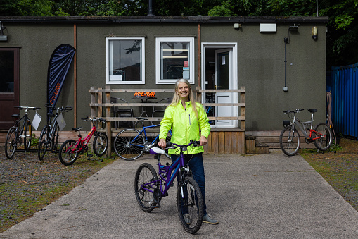 A front-view full length portrait shot of a mature woman working at a community cycle hub bike store, she is standing with a repaired bike outside the store front in Hexham, England. The organisation is a mental health charity that connects people via cycling. The cycling hub encourages, fitness, education with a view to employment, friendship, mental well-being, mental health, volunteering, leadership skills, and community bike rides.