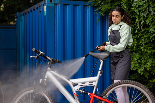 A three quarter length shot of a young female employee working at a community cycle hub bike store, she is high pressure cleaning a bike so it's ready to be hired or bought from the store in Hexham, England. The organisation is a mental health charity that connects people via cycling. The cycling hub encourages, fitness, education with a view to employment, friendship, mental well-being, mental health, volunteering, leadership skills, and community bike rides.