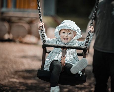 Toddler laughing at the camera as she is pushed on a swing by her dad in a play park