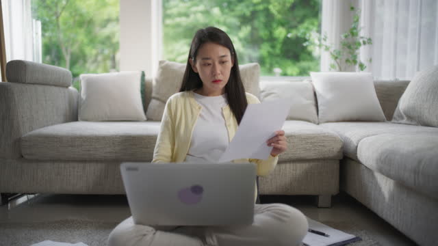 Young Beautiful Asian Female Browsing Internet, Using Laptop Computer at Home, Booking Places of Interest and Hotels for Her Vacation Trip While Sitting on a Floor in the Living Room
