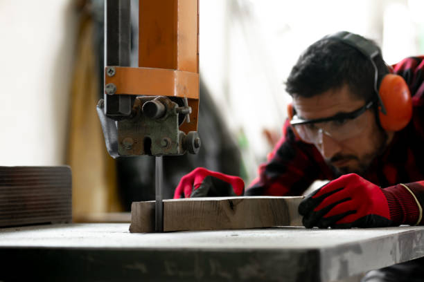 A young carpenter in a red checkered shirt and personal protective equipment processes wood in the workshop with a table band saw