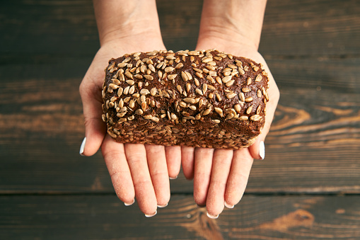 Loaf of fresh made rye multigrain bread in woman hands on wooden table background. Bakery, food concept