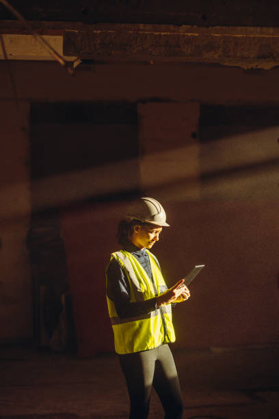 Female Worker indoors on Construction Site with sun beam falling on dust with PPE on tablet