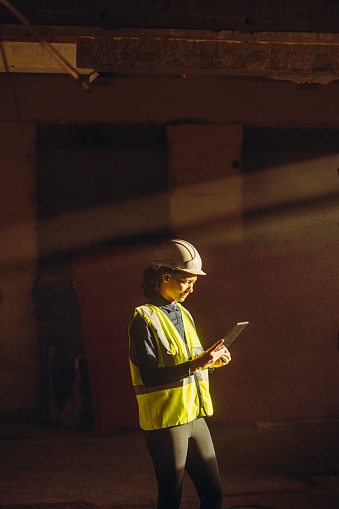 Female Worker with the correct Personal Protective Equipment working on tablet on Construction Site looking at plans.  The sun is falling on her from outside and is making a sun beam in the dust filled indoors room where she is standing examining the building process to see if the plans on her digital device is being followed.  She is giving a progress report online wirelessly back to the cloud and office.