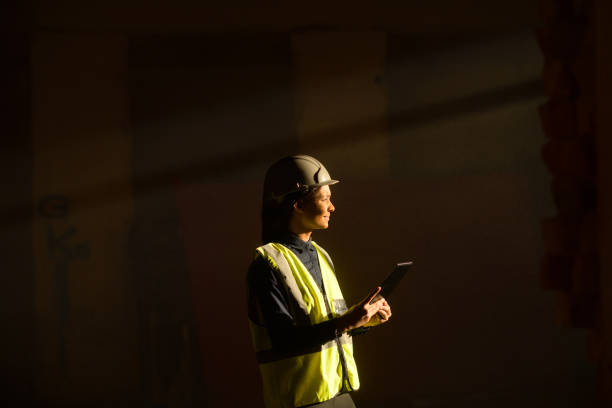 istock Female Engineer indoors on Construction Site with sun beam falling on dust with PPE on tablet 1498704939
