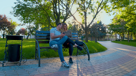 Couple is sitting on a bench in the park, both looking at phone and laughing