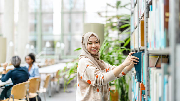 Asian muslim female student picking book from bookshelf in library