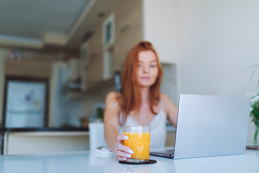 Young adult woman working from home using laptop and drinking fresh orange juice