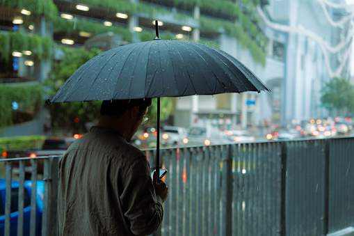 A young man using a mobile phone to hail a taxi and holds an umbrella while walking outside in the rain.