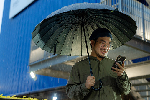 A young man using a mobile phone to hail a taxi and holds an umbrella while walking outside in the rain.