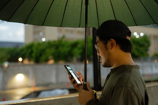 A young man using a mobile phone to hail a taxi and holds an umbrella while walking outside in the rain.