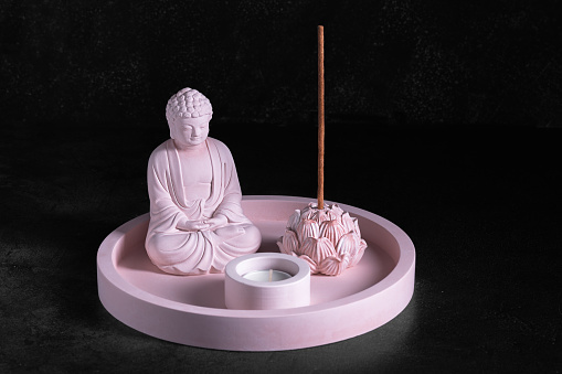 Gypsum products for meditation, relaxation, aromatherapy on a black background. Statuette of Buddha, lotus flower-shaped stand with incense stick, candlestick on a tray, low key, selective focus.