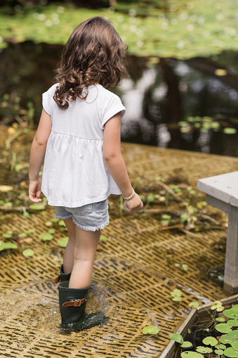 Lifestyle Candid Moment of a 4-Year-Old Girl Wearing a White T-Shirt, Deium Flowers, & Dark Green Rain Boots While Walking in a West Palm Beach, FL Garden on a Hot Summer Day