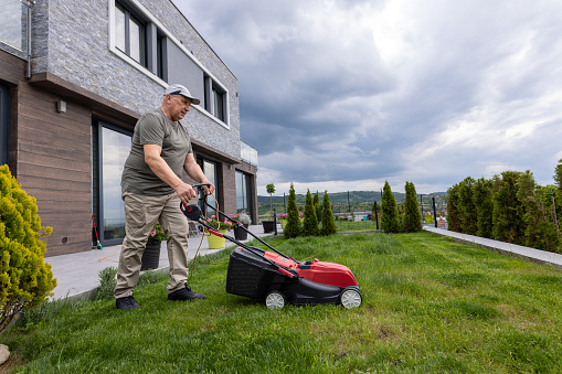 view of man in casual clothes mows lawn with lawn mower at backyard of his house. Husband takes care of garden on spring cloudy day. Modern gasoline garden equipment. Landscaping work.