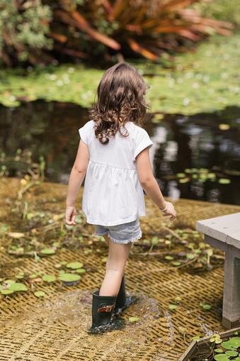 Lifestyle Candid Moment of a 4-Year-Old Girl Wearing a White T-Shirt, Deium Flowers, & Dark Green Rain Boots While Walking in a West Palm Beach, FL Garden on a Hot Summer Day