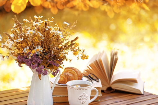 Bouquet of flowers, croissant, cup of tea or coffee, books on table in autumn garden. Rest in garden, reading books, breakfast, vacations in nature concept. Autumn time in garden