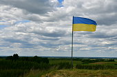 A close up on an Ukrainian flag on a pole located on the top of a tall hill with the view of numerous forest, moors, fields, pasturelands and other flora seen on a sunny summer day