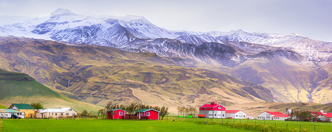 Towering snow capped mountains with cascading waterfalls rise behind Icelandic farmlands on Iceland's south coast.