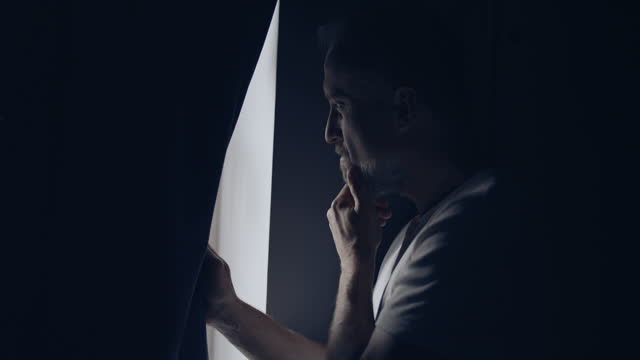 Anxious man alone in a dark room. Looking out the window with worry and touching face in distress