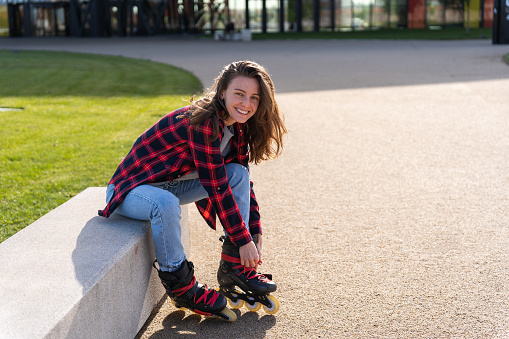 Blonde young caucasian woman putting on the rollerblade inline skates tying laces while sitting on a park and looking at the camera on a sunny day