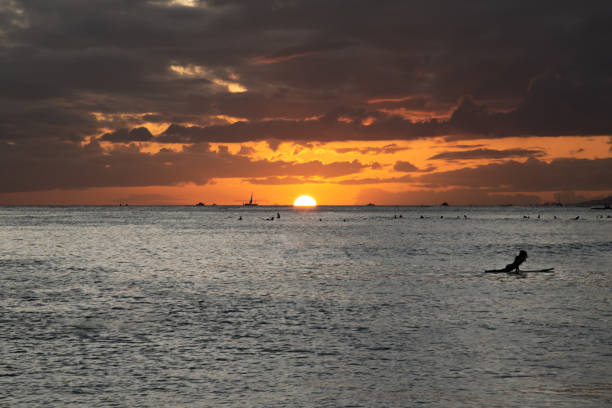 Incredible sunset over Waikiki beach on Oahu, Hawaii with sun shining bright over horizon with dramatic clouds at golden hour. People swimming and surfing in distant view.