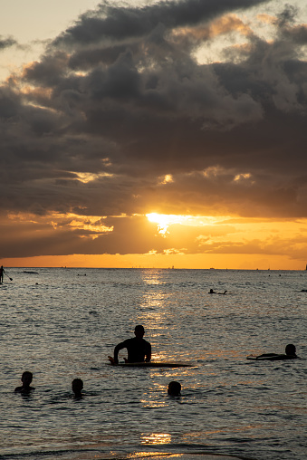Incredible sunset over Waikiki beach on Oahu, Hawaii with sun shining bright over horizon with dramatic clouds at golden hour. People swimming and surfing in distant view.