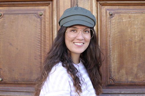 A young fashionable woman in her twenties is wearing a green beret ,retro golden eyeglasses and a pink and white striped shirt. She is posing in urban background.