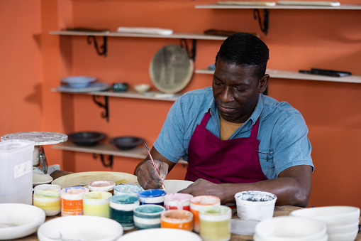 African-american man ceramist painting new crafted plate with colors and brushes.