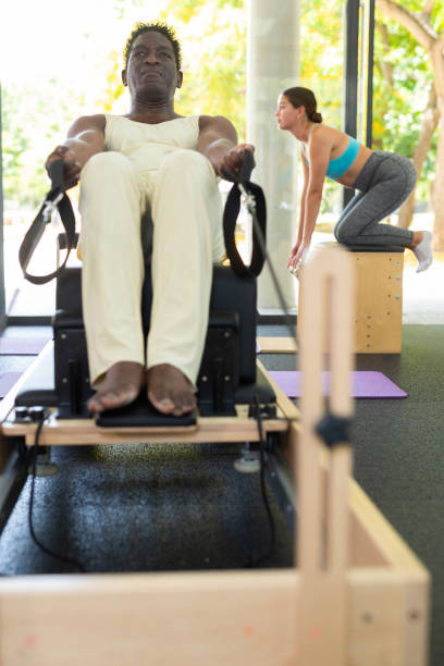Man doing exercises on pilates reformer African-american man doing exercises on pilates reformer during training in gym. Caucasian woman training on pilates chair in background. kenyan man stock pictures, royalty-free photos & images