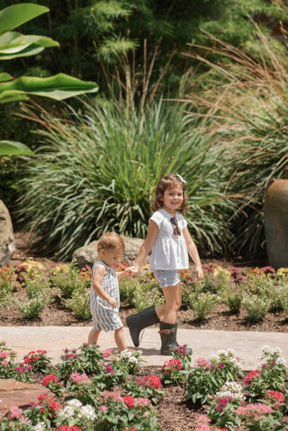 Lifestyle Candid Moment of a Young Brother & Sister Walking Together Holding Hands Wearing Sage Green & White Casual Summer Wear in a West Palm Beach, FL Garden, Cuban-American 4-Year-Old Toddler Girl & 1-Year-Old Baby Boy Both with Brown Hair estilo de vida momento sincero de un hermano y hermana jóvenes caminando juntos tomados de la mano vistiendo ropa casual de verano verde y blanco salvia en un jardín de west palm beach, fl, niña cubana-americana de 4 años y bebé de 1 año, ambos con c