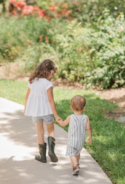 Lifestyle Candid Moment of a Young Brother & Sister Walking Together Holding Hands Wearing Sage Green & White Casual Summer Wear in a West Palm Beach, FL Garden, Cuban-American 4-Year-Old Toddler Girl & 1-Year-Old Baby Boy Both with Brown Hair estilo de vida momento sincero de un hermano y hermana jóvenes caminando juntos tomados de la mano vistiendo ropa casual de verano verde y blanco salvia en un jardín de west palm beach, fl, niña cubana-americana de 4 años y bebé de 1 año, ambos con c