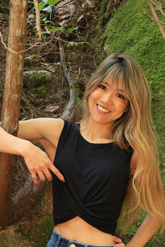 A closeup of a Japanese model with one arm around a young tree in a forest. She is wearing long, blond, straight hair, makeup, smiling, a ring, a black sleeveless, crop top and jeans.