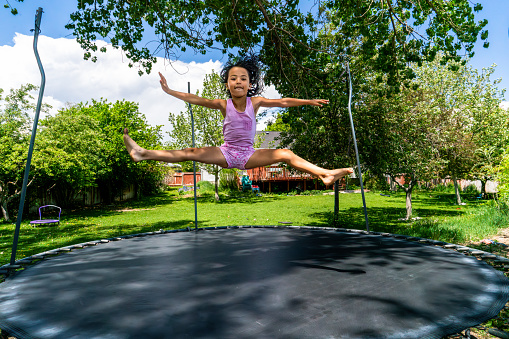 In the sun-drenched backyard, a little girl of around 6 years old leaps joyfully on the trampoline. Her long dark hair cascades as she bounces on the vibrant pink surface, her pink tank top and floral shorts mirroring her infectious happiness. The expansive yard is adorned with lush green grass and encircled by trees, providing a picturesque backdrop. Behind the trees, a cozy home with a wooden deck peeks through, blending seamlessly with the natural surroundings. The warm summer day envelops her, and she revels in the blue skies above, her beaming smile illuminating the scene.