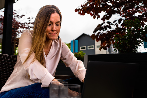 A young businesswoman exudes a professional yet relaxed aura as she works at an outdoor café. Dressed in casual business attire, she combines a light-colored blouse with comfortable jeans. Seated at a coffee table, her laptop becomes her productive companion, enabling her to tackle tasks efficiently. Above her, a pergola stands adorned with cascading pink flowers, creating a picturesque canopy. The ambiance is serene, with a gentle breeze rustling through nearby trees. With her focused gaze and poised demeanor, she seamlessly blends work and leisure in this tranquil setting, finding inspiration in the harmony of nature and her own professional aspirations.
