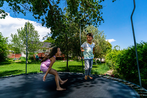 In the embrace of a large backyard, a trampoline springs to life, carrying the laughter of a little girl and boy. Framed by verdant green grass and a gentle line of trees, a home with a wooden deck adds charm to the scene. The summer day unfolds with azure skies, casting a warm glow on their joyous play. The little boy, adorned in a white t-shirt with vibrant green and yellow stripes and jeans displaying knee holes, radiates a contagious smile. By his side, the little girl, her long dark hair cascading, wears a pink tank top and delightful floral shorts, mirroring his delight. Their laughter rings out as they bounce higher, reveling in the magical harmony of the moment.