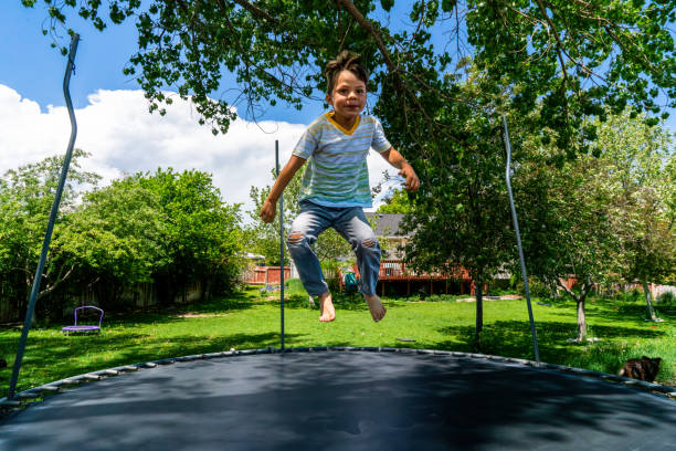 Under the warm summer sun and a sky painted in shades of blue, a little boy of around 5 years old leaps and bounds on a trampoline nestled in a sprawling backyard. The lush green grass serves as his bouncy playground, framed by majestic trees that stand tall, forming a natural perimeter. Beyond them, a welcoming home with a wooden deck peeks out, almost hidden amidst the verdant foliage. With a contagious smile on his face, the boy wears a white t-shirt adorned with playful green and yellow stripes, paired with jeans that bear the marks of childhood adventures with worn-out knees. In this idyllic scene, joy fills the air as the boy's jumps and laughter create moments of pure delight.