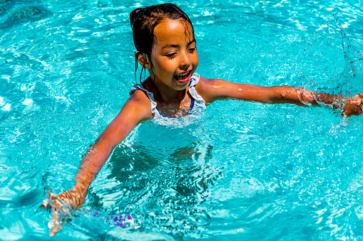On a balmy summer day, a delightful little girl, around 7 years old, gleefully splashes and plays in a pool. Her tan skin glistens under the sun's rays as her dark hair, pulled back into a ponytail, sways with each splash. With a diving baton in her hand, she gracefully dives and emerges from the water, her joyous laughter filling the air.