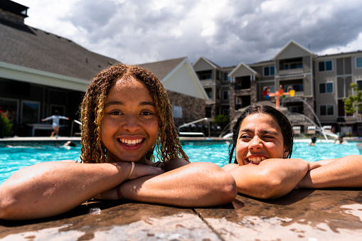 Against the backdrop of a sun-kissed pool, two multi-racial girls, aged 13 to 15, exude joy and friendship. One, with her curly blond-dyed shoulder-length hair, and the other, sporting waist-length dark hair, share heartwarming smiles and infectious laughter, capturing the essence of carefree summertime moments.