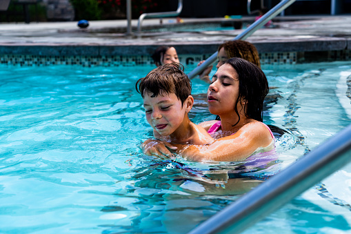 On a scorching summer day, four delightful siblings, a mix of ethnicities, revel in the cool waters of a pool. The oldest girl, a 14-year-old African American with blond-dyed shoulder-length hair, exudes confidence. Her companion, a multi-ethnic girl, proudly flaunts her long black hair. The third sibling, a young multi-racial girl of around 7, radiates innocence, her dark hair tied up in a short ponytail. The youngest, an energetic 5-year-old boy, splashes about, his laughter echoing in the air.