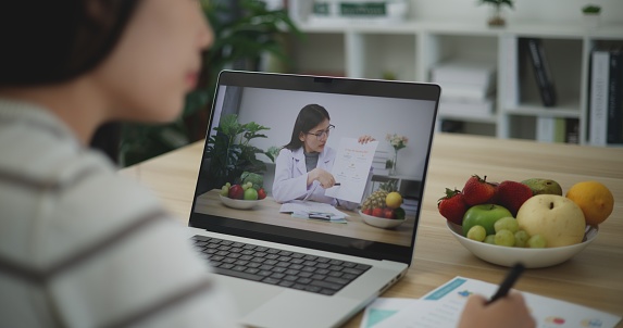 Rear view, Young asian woman on video call with female nutritionist to discuss health care plan through laptop computer, correct nutrition and diet concept