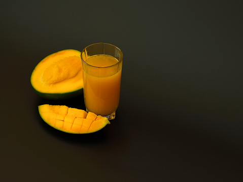 Mango juice in a tall glass on a black background, next to pieces of ripe exotic fruit. Close-up.