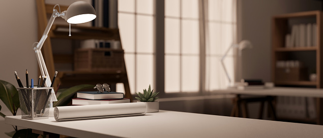 Close-up image of an architect or engineer's office desk in a modern office room with a roll of blueprints, pencils in a pencil stand, books, a table lamp, and copy space. 3d render, 3d illustration