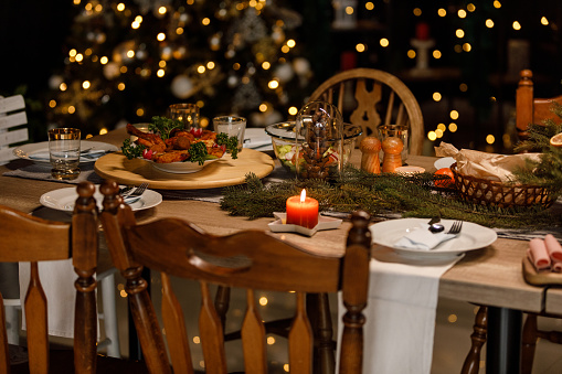 Selective focus shot of glistening Christmas lights in the background, behind a rustic dining table set for a Christmas dinner party.