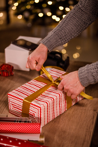 Close up shot of unrecognizable young man standing over a table and tying a bow on top of a Christmas gift that he wrapped.