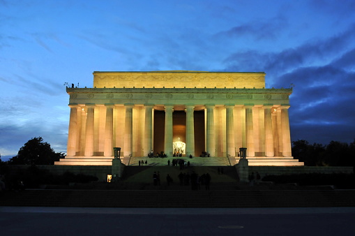 Tourists visit the Lincoln Memorial in Washington, D.C. on an autumn night. Long exposure wide shot from the east side of the building. The lights of a passing airplane are seen in the sky.