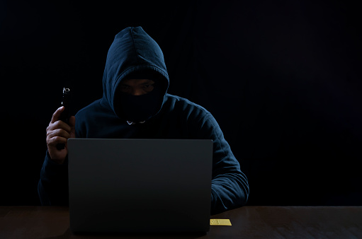Scene of hacker wearing thief hoodie with gun is hacking computer system on laptop, cyber crime concept